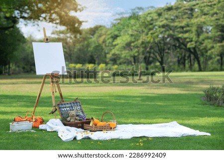 Picnic basket, fruits basket on picnic cloth, drawing and easel with frame on lawn outside in summer park, summer picnic background concept