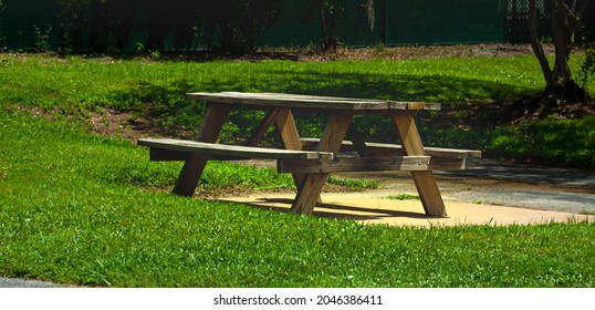 A Picnic Bench Surrounded By Grass And The Sun Is Beaming Down On The Table.  The Bench Is Made Of Wood And Seats 3 People On Each Side.