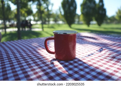 Picnic Concept, Shiny Beautiful Weather Picnic Day, Red Coffee Mug On Picnic Table, Table Covered With Square Layout. Park And Outdoor Picnic Idea, No People.