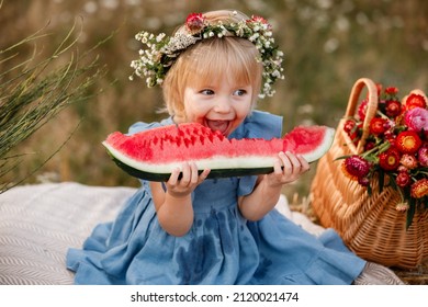 picnic with family. cute little girl eating big piece of watermelon on straw stack in summertime in the park. Adorable child wearing in flowers wreath on head and blue dress
