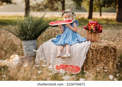 picnic with family. cute little girl eating big piece of watermelon on straw stack in summertime in the park. Adorable child wearing in flowers wreath on head and blue dress