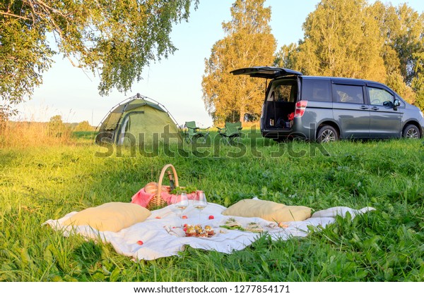 Picnic in the forest, tent and camp chairs, gray\
minibus with open door, blanket, wicker basket, wine glasses,\
snacks, fruit, bread, pillows. Concept of romantic camp date nature\
in sunny day