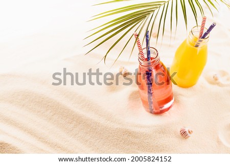 Picnic with fruit cocktails on tropical beach, vacation background - fresh cold pink and yellow drinks with straw in glass bottles, green palm leaf in golden sunlight on white sand, copy space.