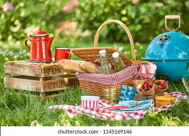 Picnic Hamper And BBQ In The Shade Of A Tree In A Lush Green Garden With Fresh Berries, Bread, Soft Drinks And Colorful Red Enamel Coffee Can On A Checkered Tablecloth