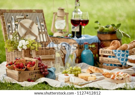 Picnic hamper surrounded by delicious fresh food with berries, fruit, cheese, bread rolls, wraps glasses of red wine, chilled infused water, pastries for dessert and a vintage lantern on green grass