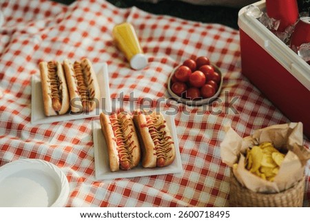 Picnic with hot dogs, chips and cold drinks in cooler bag on red tablecloth in a park.