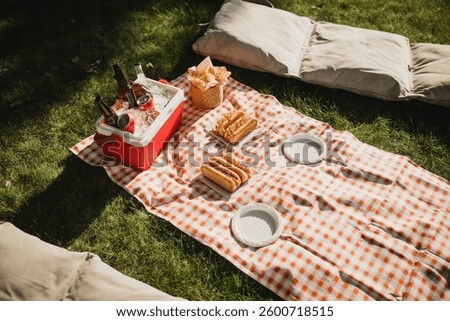 Picnic with hot dogs, chips and cold drinks in cooler bag on red tablecloth in a park.