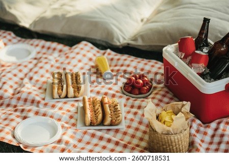 Picnic with hot dogs, chips and cold drinks in cooler bag on red tablecloth in a park.