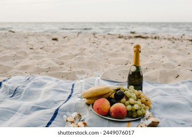 Picnic on beach on summer day. Champagne, sweets and glasses. A plate of fruit on a blanket: grapes, bananas, peaches, and plums. Romantic date in nature for spending time together on the sand sea. - Powered by Shutterstock