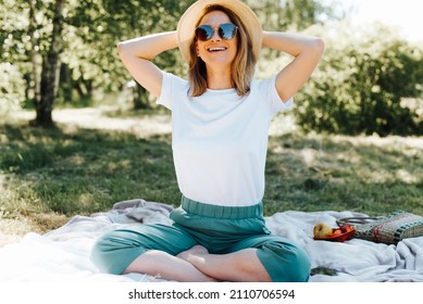 Picnic, rest, relaxa. Laughing caucasian young woman in straw hat and glasses sitting in lotus position on blanket, outdoors. Pretty freckled happy girl enjoying leisure time on summer day.