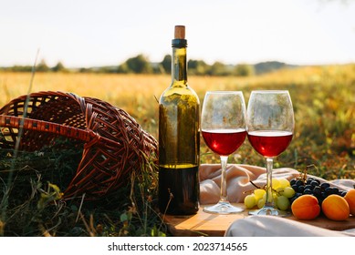 Picnic, romantic evening in nature concept. Bottle of red wine, glasses with drink, grapes, peaches on wooden board, wicker picnic basket on sunset background - Shutterstock ID 2023714565