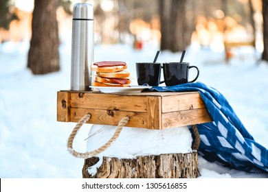 Picnic With Sandwiches And Thermos With Tea Or Coffee In Winter Park Or Forest. Two Cup For A Pair Of Lovers Or Tourists On A Wooden Box On The Snow. Toast Bread, Cheese, Ham. Romantic Weekend Party