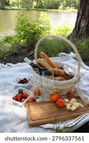 Picnic setup featuring a basket with bread, cheese, and fruits on a blanket near a tranquil lake with greenery and trees in the background