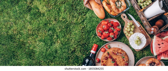 Picnic Straw Basket With Healthy Food, Accessories. Summer Picnic With Cake, Fruits, Cheese, Wine And Snacks On Plaid Over Green Background. Top View. Copy Space. Summer Family Lunch. Romantic Picnic.