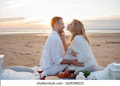 Picnic at sunset. A man and a pregnant woman sit on a bedspread, among fruits and pillows, on the beach, hugging, kissing against the backdrop of low tide and a wide coastal strip.