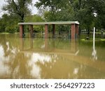 Picnic tables nand pavillion are flooded at the park. Trees are in the background and flood water is in the foreground.