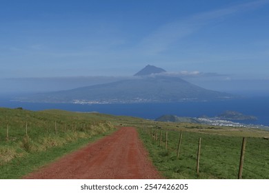 Pico Volcano on the Azores viewed from Faial Island on a sunny spring day. A red-brown dirt road winds through green meadows, offering a stunning view of the volcano on the neighboring island.   - Powered by Shutterstock