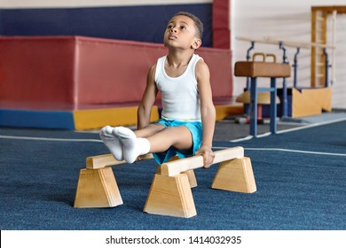 Picture of adorable dark skinned little gymnast competing on parallel bars. Hardworking talented African child exercising at gym, doing acrobatic moves, demonstrating strength, agility and flexibility