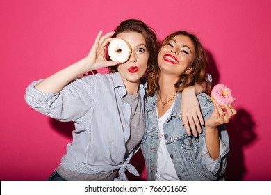 Picture Of Amazing Two Women Friends Eating Donuts Isolated Over Pink Background. Looking Camera.