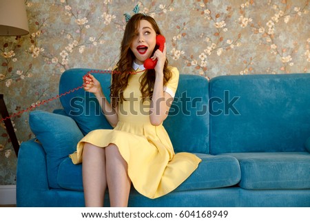 Picture of amazing young pin-up lady with red lipstick sitting on sofa at home. Looking aside while talking by phone.