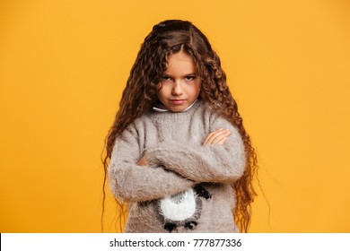 Picture Of Angry Little Girl Child Standing Isolated Over Yellow Background. Looking Camera With Arms Crossed.