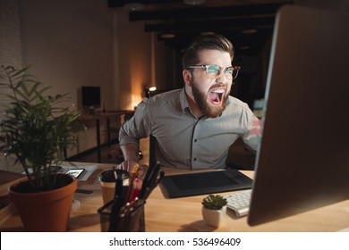 Picture Of Angry Web Designer Dressed In Shirt And Wearing Eyeglasses Working Late At Night And Looking At Computer While Screaming.