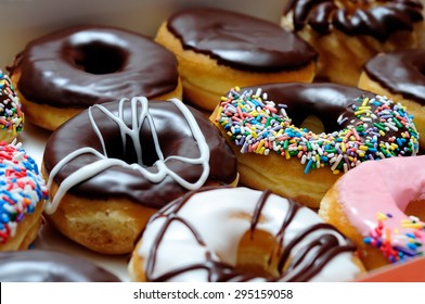 Picture of assorted donuts in a box with chocolate frosted, pink glazed and sprinkles donuts.