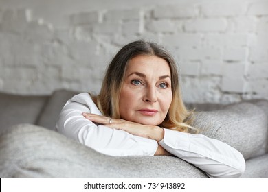 Picture of attractive middle aged Caucasian woman with long straight hair resting on grey comfortable sofa, having sad unhappy expression, feeling bored or lonely. People, lifestyle and age concept
