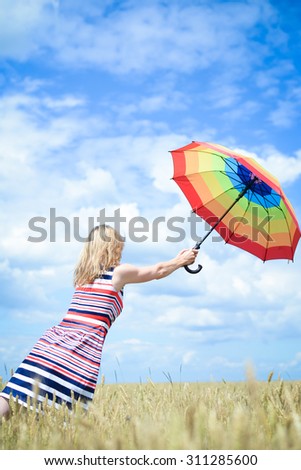 Picture of beautiful blond woman flying away in the wheat field. Ladi in multi-coloured stripped dress holding umbrella.
