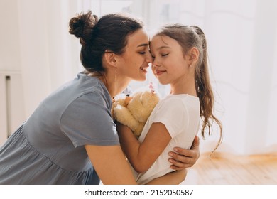 Picture of beautiful caring young mother hugging her cute little daughter tight, holding teddy bear with eyes closed, embracing her child gently, showing her love, protection and admiration