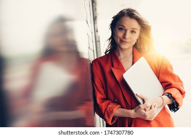 Picture Of Beautiful Confident Young Businesswoman Lean To Wall And Pose On Camera. Girl Hold Laptop In Hands. Remote Work Outside Of Office. Sun Shines Behind Woman. Stylish Trendy Girl On Picture