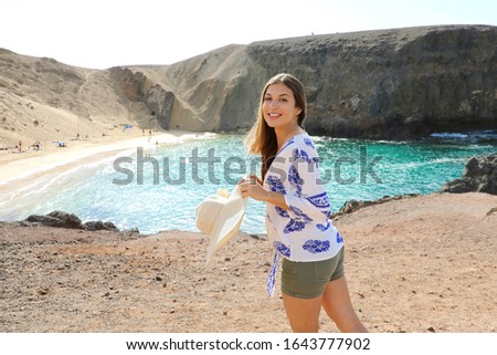 Picture of beautiful tourist woman with Playa Papagayo landscape on background. Happy girl with hat in her hand on Lanzarote island with Playa Papagayo beach on Canary Islands, Spain.