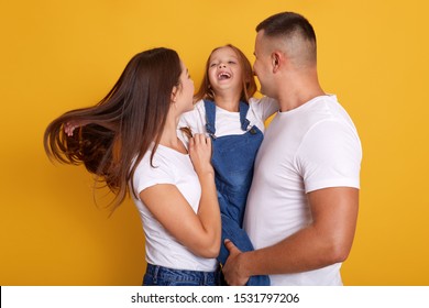 Picture Of Beautiful Young Family Hugging And Looking Each Other, Laughing While Standing Against Yellow Studio Background, Having Fun Together, Looks Happy, Expresses Gladness. Family Concept.