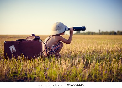 Picture of boy wearing pith helmet and plaid romper looking in spyglass in wheat field. Little explorer with camerabag and old suitcase on sunny countryside background. - Shutterstock ID 314554445