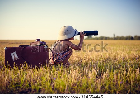 Picture of boy wearing pith helmet and plaid romper looking in spyglass in wheat field. Little explorer with camerabag and old suitcase on sunny countryside background.