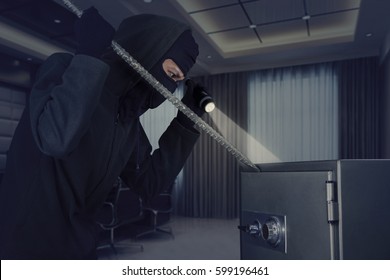 Picture Of Burglar Stealing A Safe Deposit Box Using A Crowbar While Holding A Flashlight In The Office