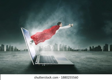Picture of Caucasian businessman wearing red cape while flying out of laptop screen with cloudy city background