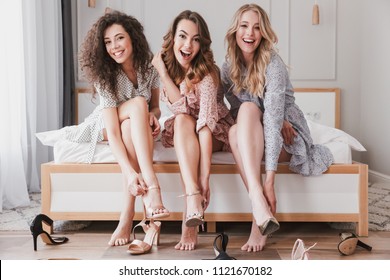 Picture of caucasian stylish girls 20s wearing dresses trying on different summer shoes or high heels during bachelorette party in cozy bedroom