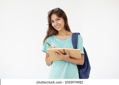 Picture Of Cheerful Positive Hispanic College Girl With Eyeglasses On Her Head And Bag On One Shoulder Posing In Studio, Smiling Happily While Writing Something Down In Her Copybook Or Diary