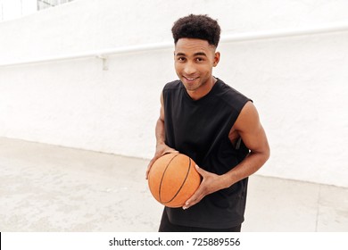 Picture of cheerful young african man walking outdoors. Looking camera play basketball.