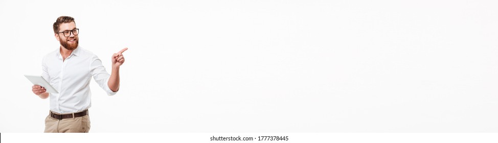 Picture of cheerful young bearded man standing near copyspace over white wall background isolated. Looking aside using tablet computer pointing.