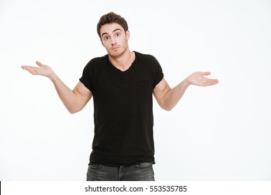 Picture of confused young man dressed in black t-shirt standing over white background gesturing with hands.