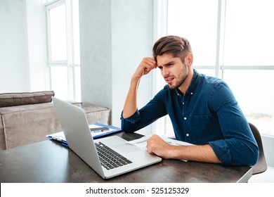 Picture Of Confused Young Man Sitting Near Laptop And Holding Head With His Hand. Looking At Laptop.