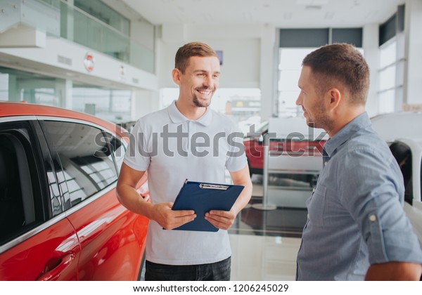 Picture of customer and seller\
stand together in front of beautiful red car. They look at each\
other and smile. Seller holds plastic tablet. They are on\
salon.