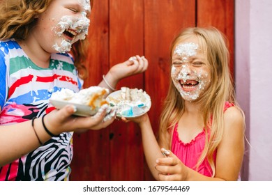 Picture of cute caucasian girl with long fair hair and in pink dress with her older chubby sister play and smear the cake across each other's faces against the wooden wall in village