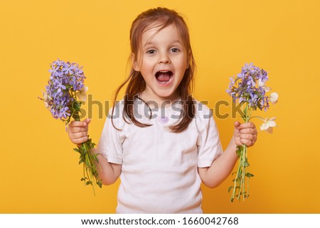 Picture of emotional playful little girl opening mouth and eyes widely, holding flowers in both hands, wearing white t shirt, preparing present for her mom on women day. Children and gift concept.