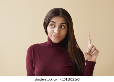 Picture Of Emotional Young African American Female Looking Away And Pointing Index Finger Up, Having Bright Idea. Beautiful Dark Skinned Woman Indicating Something On Blank Copyspace Studio Wall