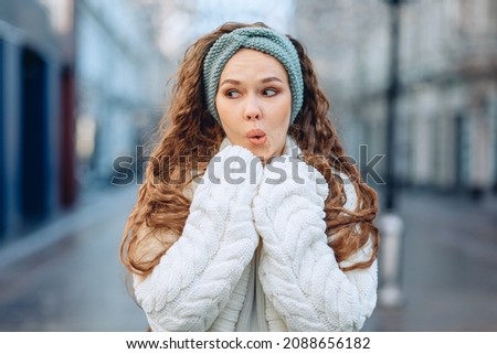 Picture with expression of state. A young girl stands posing on street in city center dressed in white knitted suit. The emotion of embarrassment and confusion. Eyes averted and mouth slightly open.