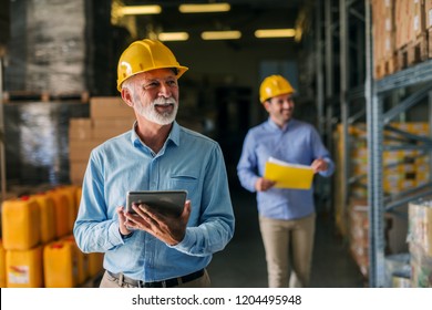 Picture of father and son walking through their warehouse with helmets on their heads. Father is holding digital tablet in his hand and looking at shelf full of boxes with smile on his face. - Powered by Shutterstock