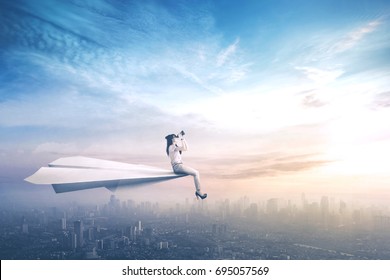 Picture of a female worker sitting on a paper aircraft while flying above a city and looking through a binoculars - Powered by Shutterstock